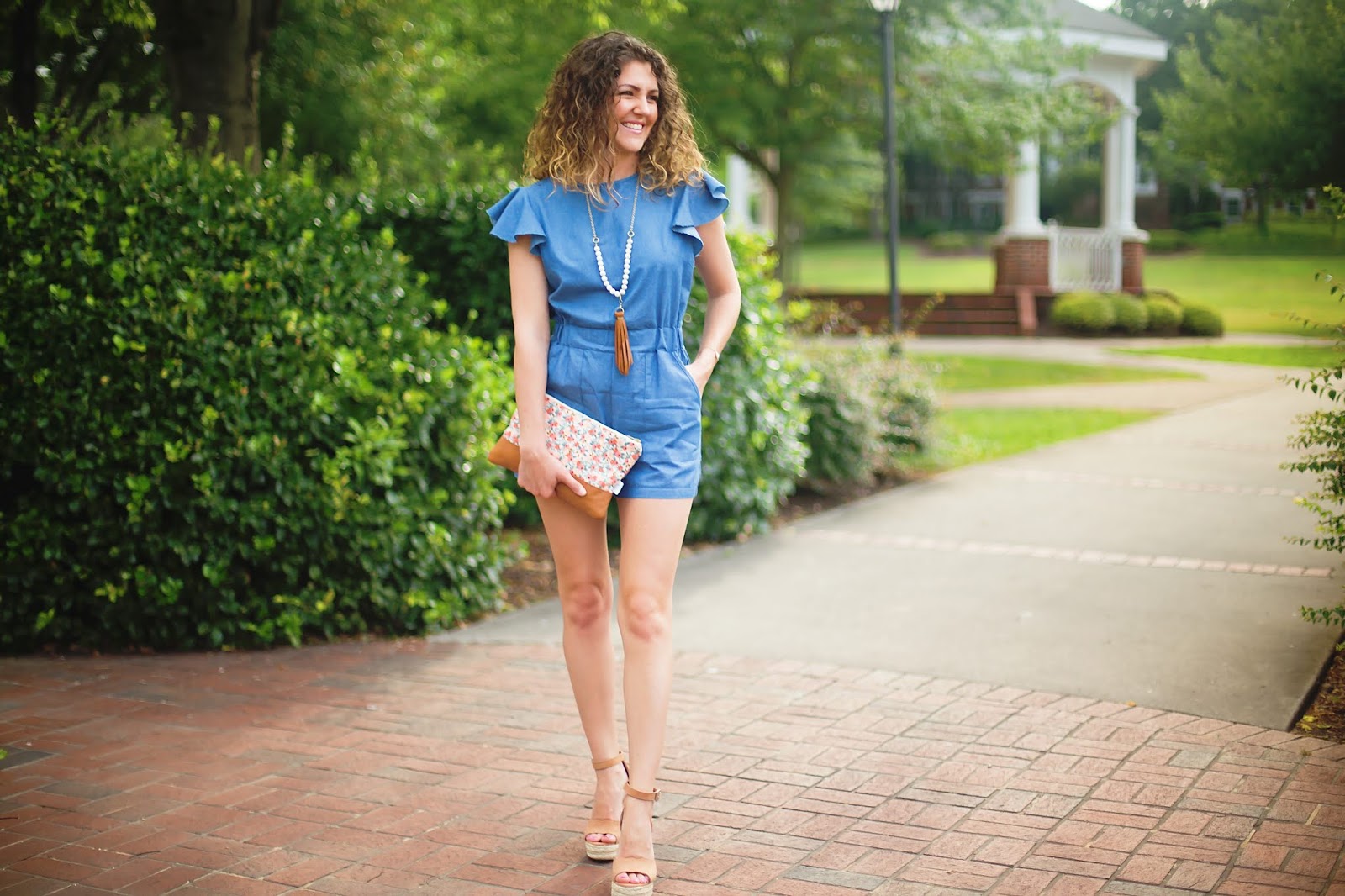 Jenn Pennell, Shaw Avenue blog, wears a denim romper from Amazon, Steve Madden scalloped wedges, floral clutch, and leather necklace.