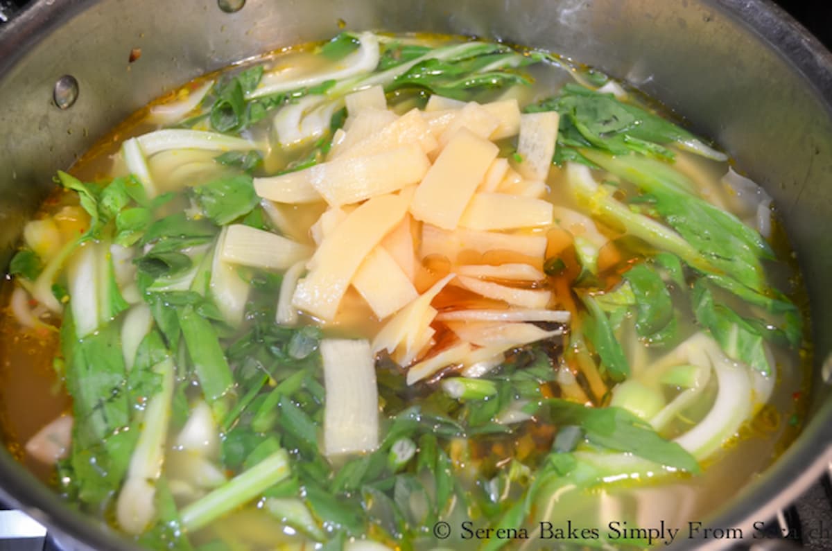 A large soup pot filled with simmering broth, wontons, sliced bok choy, sliced bamboo shoots, sliced green onions and toasted sesame seed oil.