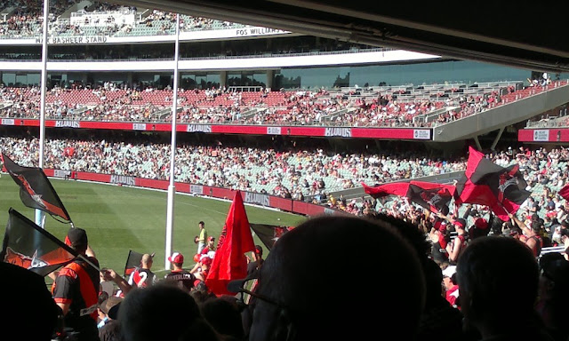 View from the bottom tier of the Adelaide Oval southern stand. One can see the four tiers of the eastern stand and the crowds.  Behind the goals the crowd is a sea of red and black guernseys and flags and banners.