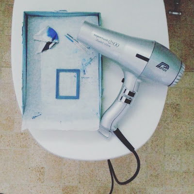 Aerial view of a toilet with the lid down. On top of the lid is a hairdryer and a box lid containing an one-twelfth scale miniature wooden picture frame, painted blue.