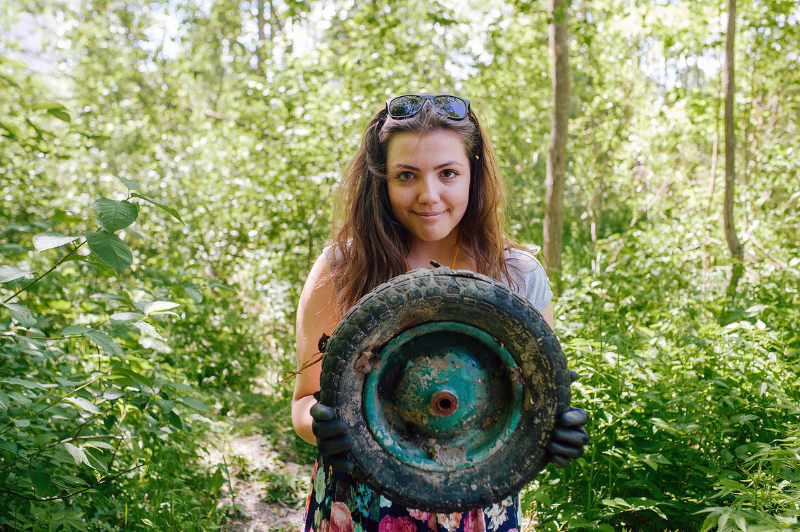 A Clean Games Baltic Cup volunteer holds up an old, rusted wheel she collected from a forest cleanup. She wears black gloves and smiles proudly, surrounded by lush greenery.