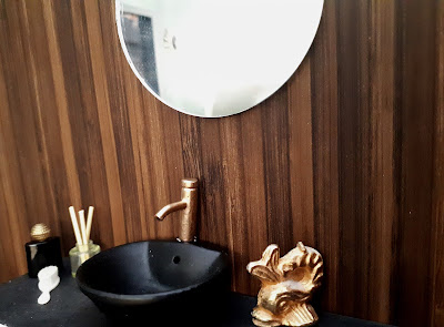 One-twelth scale modern miniature bathroom with black basic, gold tap and wood panelled wall.