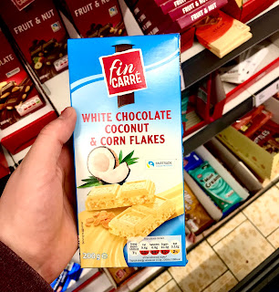 A tall rectangular white chocolate bar studded with white coconut and brown bits of cornflake in a silver metallic wrapper and a blue tall rectangular cardboard wrapper with fin carer in a red diamond with white font and white chocolate, coconut and cornflakes in white font on a bright background