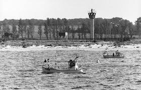 Ein Wachturm auf Land. Davor zwei Fischerboote(?), die verlassen auf dem Wasser dümpeln.