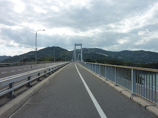 View along road deck of the Hakata Oshima bridge, first part is the girder bridge, with the suspension bridge section visible in the distance. Mountains of Oshima are visible in the backgound. On the Shimanami Kaido Bikeway