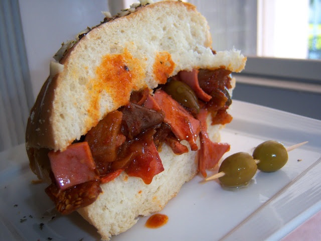 Fried Bologna and Caponata this is a fried bologna and eggplant appetizer sandwich with olives and sauce in a hard crusted Italian roll. This is on a dish by a picture window.