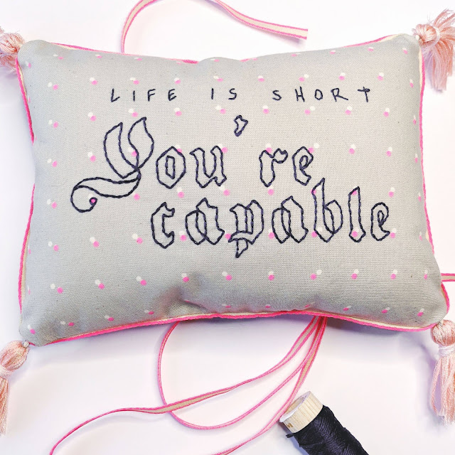 Life is short You're Capable hand embroidered on decorative pillow with tassels in the corners of the pillow and loose ribbon and thread spool next to it