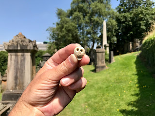 Skulferatu #44 - a photo of a small ceramic skull being held in a hand with gravestones, grass and trees in the background.  Photo by Kevin Nosferatu for The Skulferatu Project.