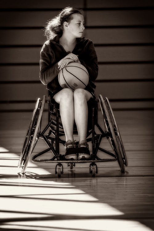 Young woman viewed face-on, sitting in a sports wheelchair with cambered wheels, clutching a basketball