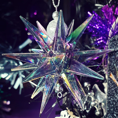 Spiked iridescent bauble A bauble in the shape of a jaggedly pointed star with many points, made of clear plastic with an iridescent rainbow finish that tends towards green and purple. The bail for suspending the bauble is two silver beads It is infront of the black Christmas tree and a dark purple wall. Bright purple tinsel is visible