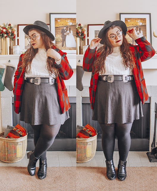 A woman with brown wavy hair wearing a black wide brim fedora, white pleated Victorian inspired blouse, a red and black buffalo plaid coat, a black short skater skirt, a black and silver western belt, black tights and black heeled ankle Chelsea boots