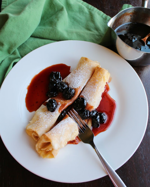 Lemon ricotta stuffed crepes are the perfect sweet bite of spring for breakfast, brunch or a light dessert. The syrup made from dried cherries is the literal cherry on top! fork cutting into stuffed crepe covered with cherry syrup. pan of syrup in background