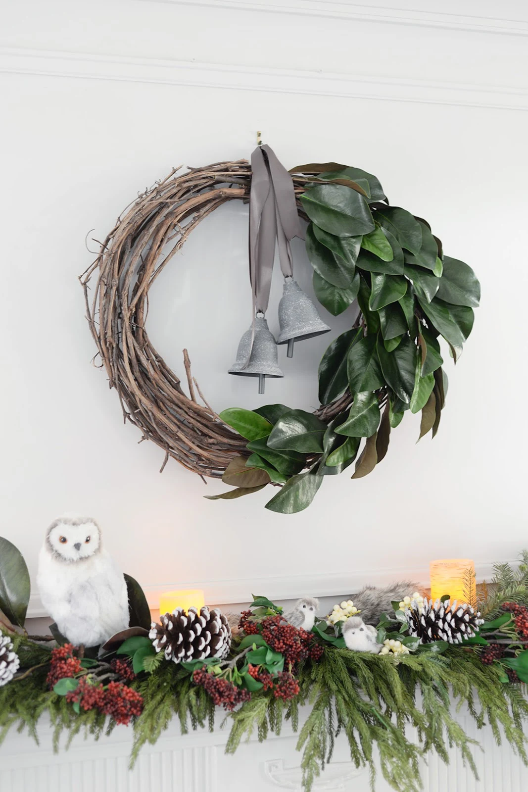 a faux christmas fireplace mantel garland with red seeded eucalyptus, a cedar garland and magnolia leaves. A vine and magnolia asymmetrical wreath with satin ribbon and metal bells. A brass chevron fireplace screen.