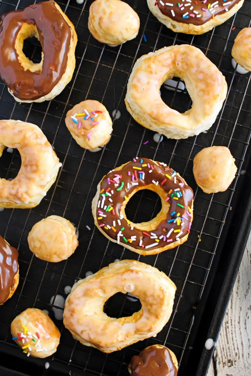An overhead photo of a variety of chocolate dipped donuts with sprinkles, glazed donuts, and donuts holes on a wire rack over a baking sheet.