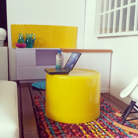 One-twelfth scale modern miniature office break-out space in bright colours with a kitchen in the background and seating and coffee tables in the foreground.