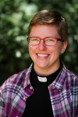 ID: Rev. Emily E. Ewing smiles with glasses and short hair, wearing a clergy collar and purple plaid shirt.