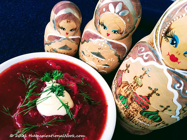 A bowl of red Russian Borscht soup with dill and sour cream surrounded by matryoshka dolls.