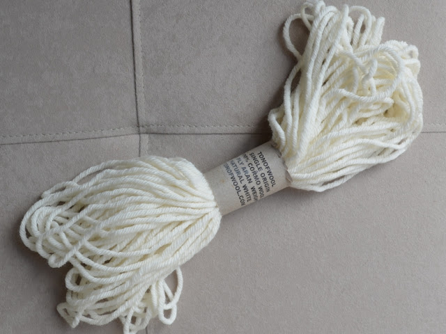A 25 gram skein of creamy, natural white, aran weight wool with a plain paper label with the details printed in bold black capitals. The text says: TONOFWOOL SINGLE ORIGIN 100% CORMO WOOL 10 PLY ARAN WEIGHT NATURAL WHITE TONOFWOOL.COM