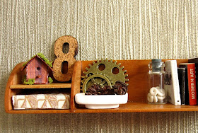 Modern dolls' house shelf  holding a small shabby bird house, a cork number 8, two cogs, a bowl full of pine cones, a jar with shell inside, holding up several books.