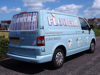 Photograph of three quarter view of an eggshell coloured Volkswagen van the back has a large window with stripes in pastel shades of pink, blue, green and yellow, Flowers in a bold white font, with a script text 'by SP ltd' underneath. Two doves on the body of the car in blue, white and pink. Vinyl lettering for the phone number and website address. On the side it has more doves and daises in a round ball shape. Flowers is in a large bold font with the same stripes running inside them.