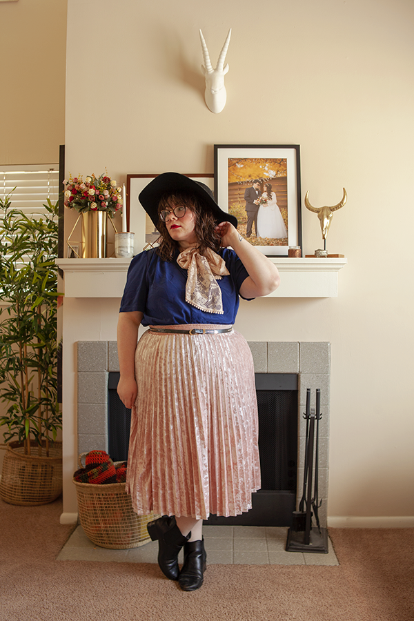 An outfit of a large black wool floppy hat, short sleeved navy blue blouse, pastel pink midi skirt and a pink lace scarf tied into an off center bow around the neck.