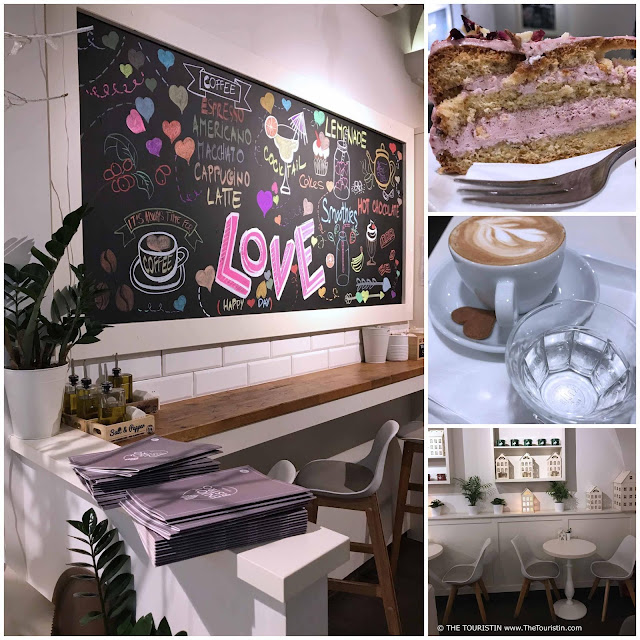 Wall mural with word art, a large piece of cake with pink filling, a cup of cappuccino served with a heart-shaped biscuit, and white chairs around a round table in front of a wall decorated with white miniature houses.