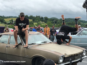 Freiburger sits on the roof of a 1972 Ford Maverick while Mike Finnegan attempts a head stand on the fender of the beater car.