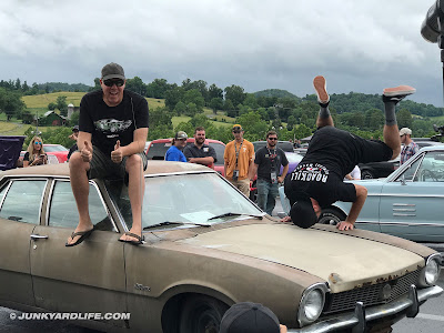 Freiburger sits on the roof of a 1972 Ford Maverick while Mike Finnegan attempts a head stand on the fender of the beater car.