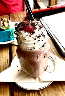 A squared cylindrical glass filled with dark brown milkshake topped with white cream and purple-brown pieces of cake  on a blue plate on a rectangular brown table on a bright background 