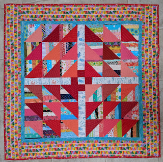 HSTs of strings pair with red, orange, and pink solid triangles to form The Square Deal block in the center of this quilt. It is surrounded by a narrow turquoise inner border and a wider print of colorful triangles.
