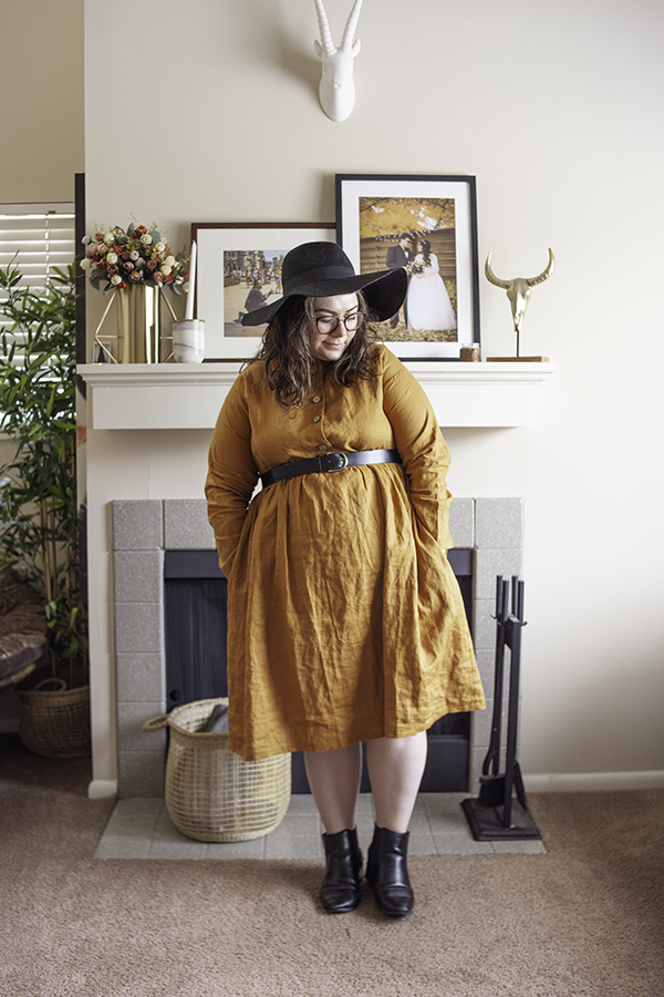 An outfit consisting of an oversized black floppy hat, a golden yellow collared button down linen dress, and black heeled Chelsea boots.