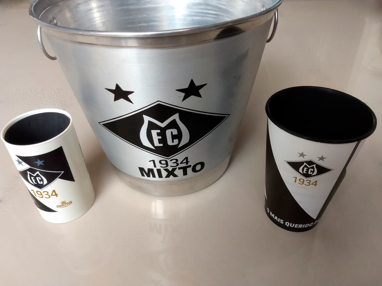 Na foto um porta lata branco do Mixto, um balde de metal inox para gelo eu copo plástico preto, produtos oficiais