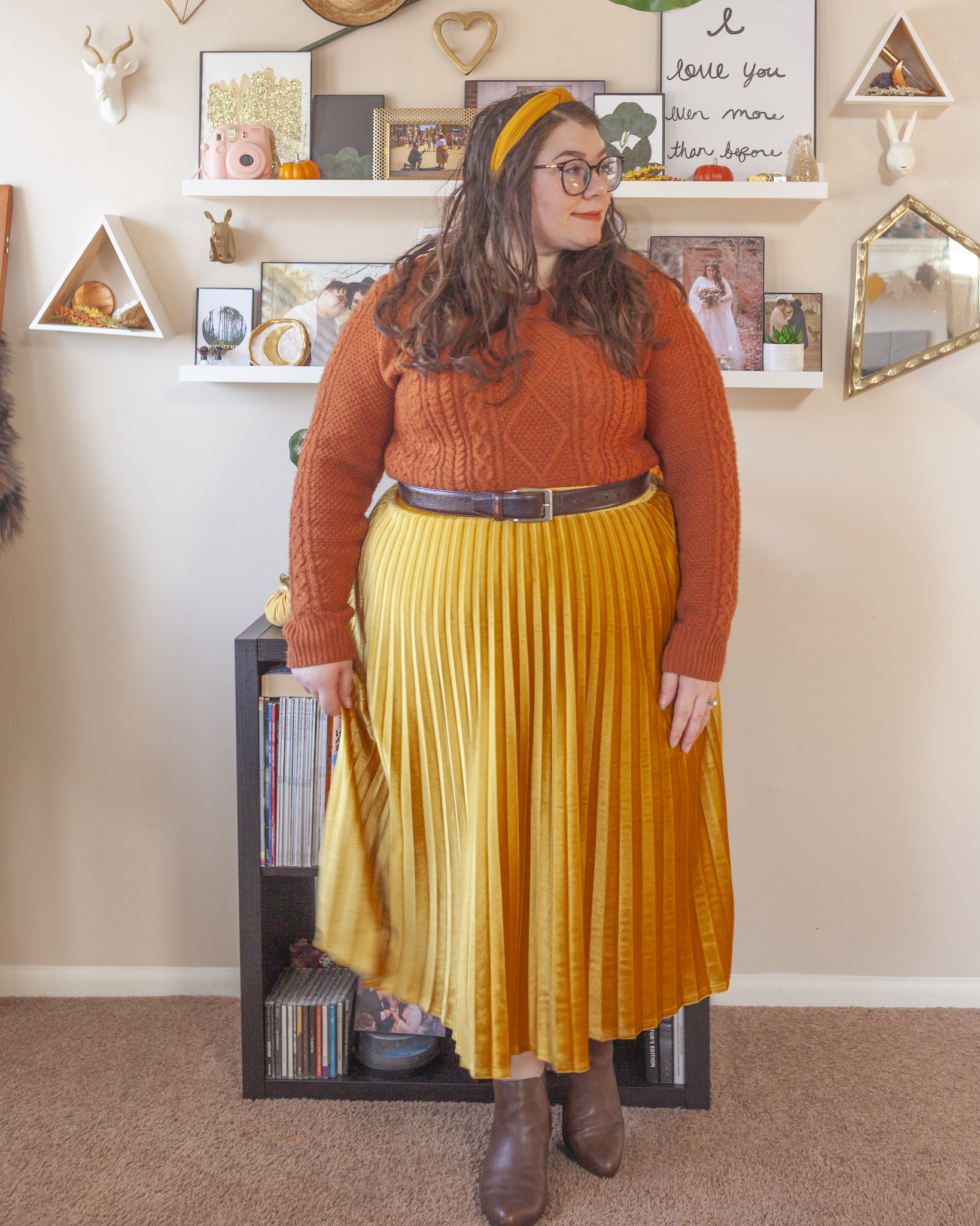 An outfit consisting of a yellow velvet knotted headband, a burnt orange crew neck sweater tucked into a mustard yellow velvet pleated midi skirt and brown Chelsea boots.