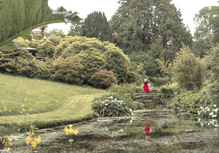 girl in red dress stands above still pond, surrounded by green trees, grass, and yellow and white flowers