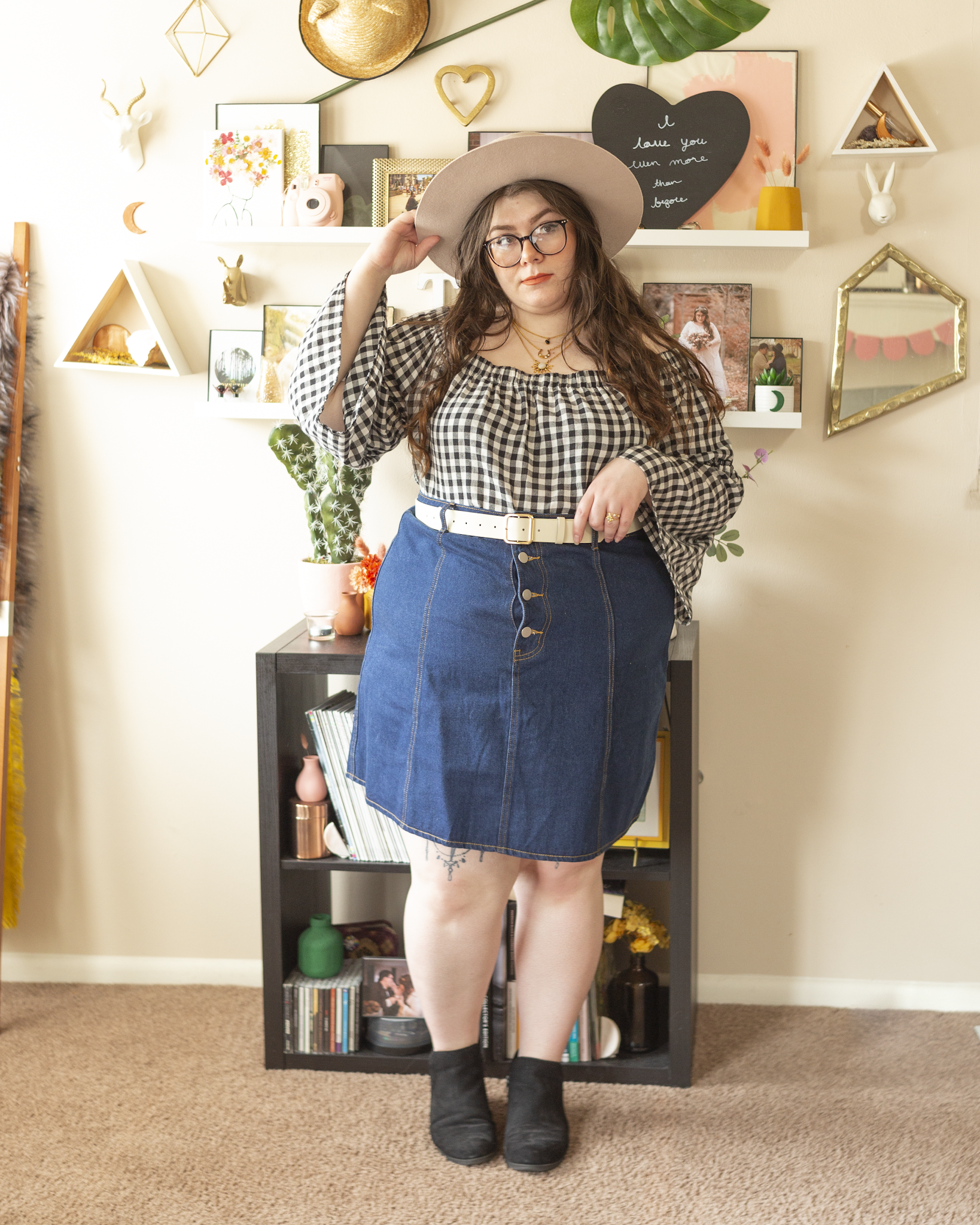 An outfit consisting of a beige wide brim porkpie hat, an off the shoulder black and white gingham blouse tucked into a an a-line denim skirt and black booties.