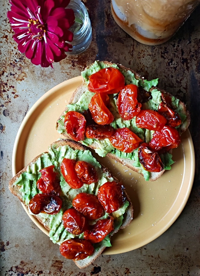 Avocado Toast With Roasted Tomatoes on a plate and a little flower and iced coffee