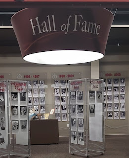 An image of a gallery in the Alberta Sports Hall of Fame. A large, central light fixture that reads "Hall of Fame" hangs above a wall hung with many portraits of Hall of Fame inductees.