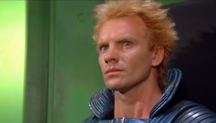 Imagem: o personagem Feyd, interpretado pelo ator e cantor Sting, com os cabelos ruivos espetados e olhos azuis em um traje cinzento com gola alta em uma sala de cor verde doentia.