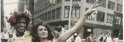 Marsha P Johnson and Sylvia Rivera, transgender icons, are visible side-by-side in a color photo. Marsha has flowers in her hair while Sylvia holds her left hand up in a peace sign.