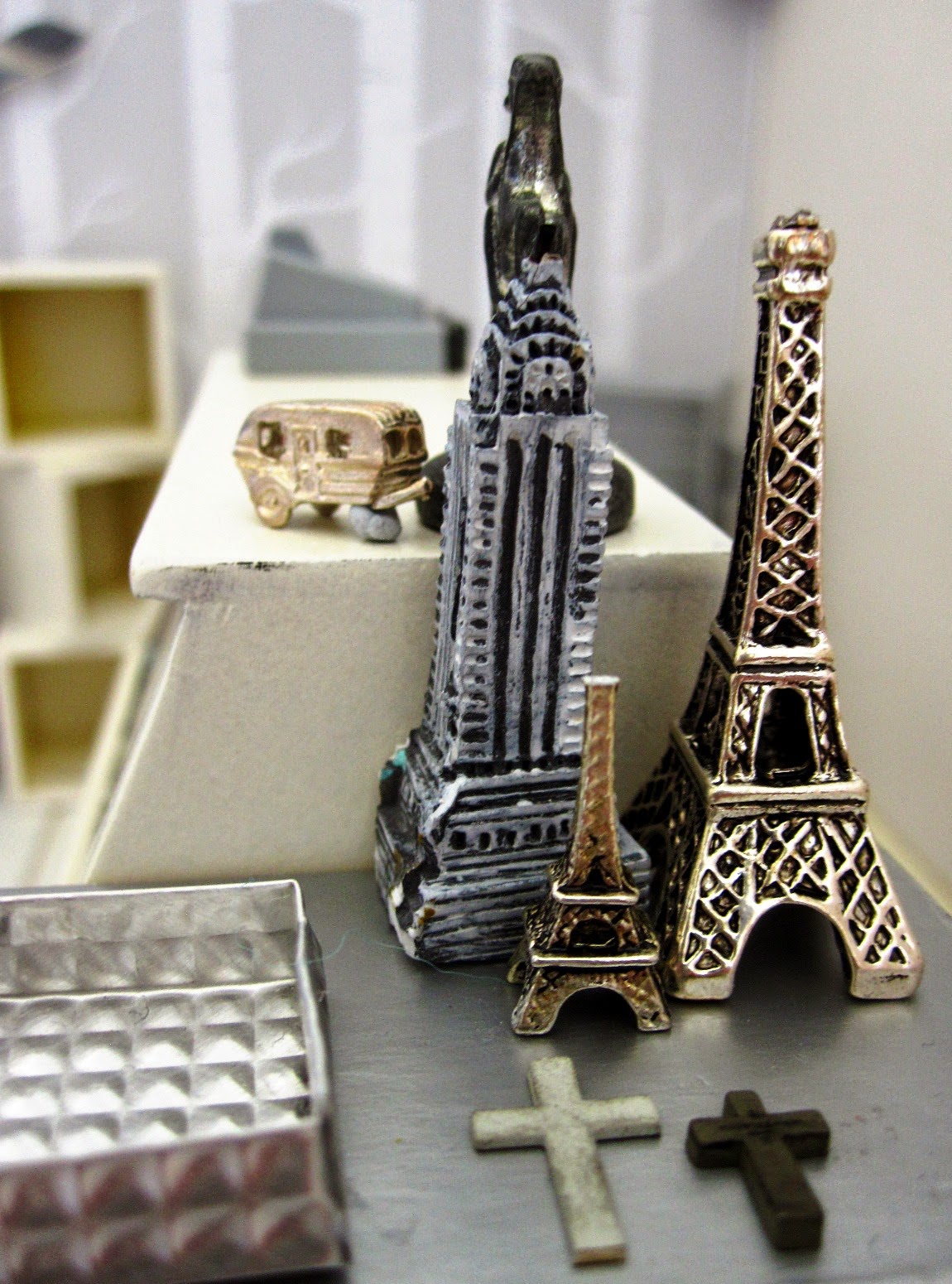 Close up of  a modern dolls' house miniature display of model buildings: an Eiffel Tower in two sizes, plus an Empire State Building. In front are two grey crosses and to the left is a grey metal tray.