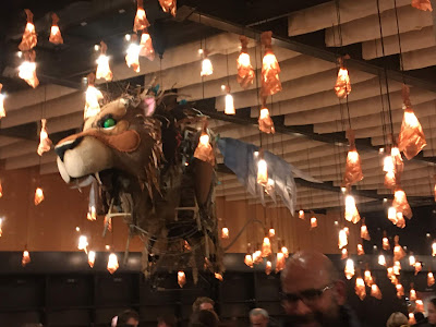 photo of a puppet of a lion's head with green eyes, suspended from the ceiling and surrounded by lights 