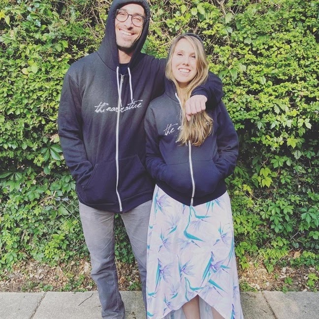 Jesse and Suzie stand in front of a green fence wearing a black hoodie that says 'the narrative' printed on it