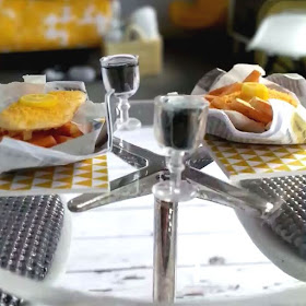One-twelfth scale miniature fish and chips for two, served in newspaper with glasses of red wine on a clear circular table.