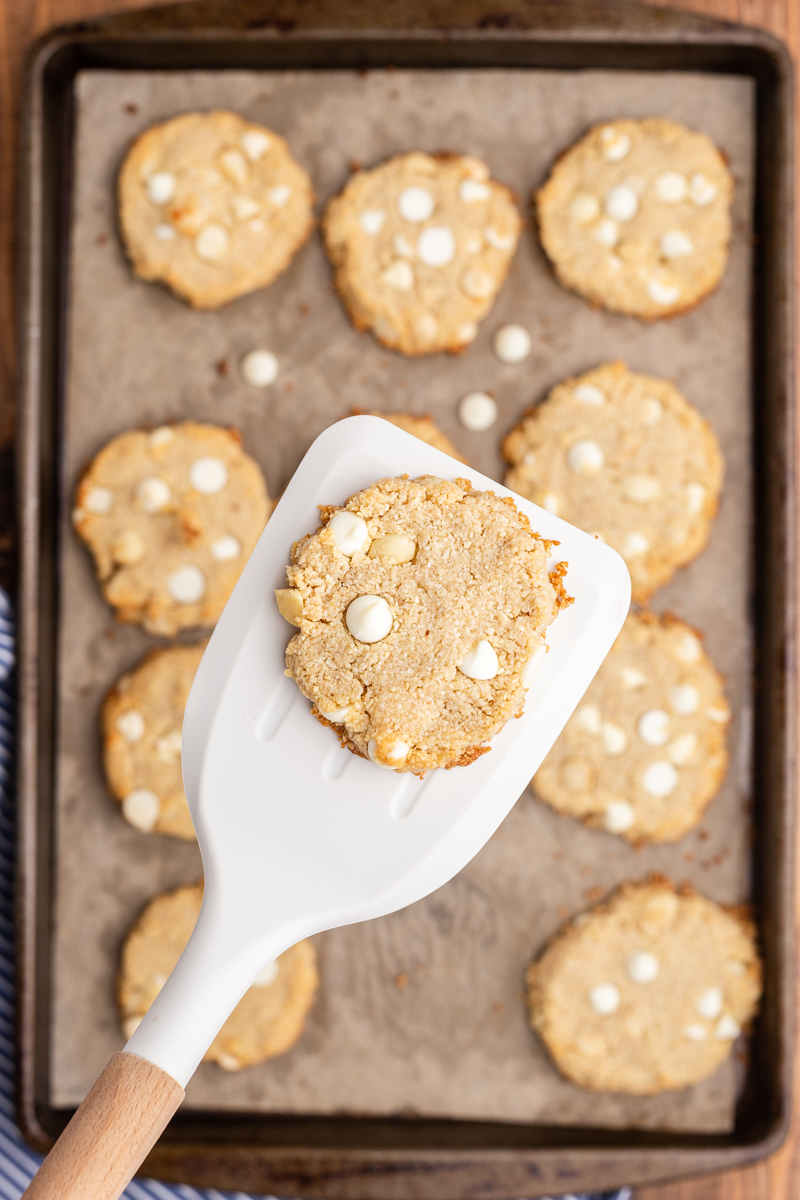 Overhead photo of Keto White Chocolate Macadamia Nut Cookies on a baking sheet with one cookie on a spatula closer to the camera.