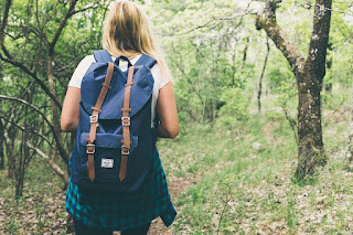 A blond girl with a blue backpack facing into the woods.
