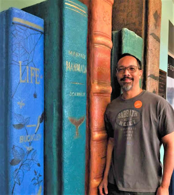 Photo of Stephen De La Vega standing next to the spines of books that are larger than life. Most prominent book is title "Life."