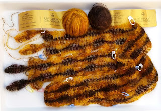 The Autumn Gold scarf is folded with two ends facing the left edge of the picture. One can see two gold leaves and two brown leaves as part of the leaf fringe. White plastic pin stitch markers indicate the pattern repeats. Above the scarf are two balls of Alchemy silk mohair yarn alongside their gold labels.