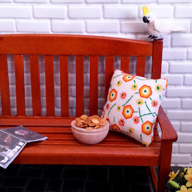 One-twelfth scale modern miniature wooden park bench in front of a white brick wall. On the bench is an IKEA catalogue, a bowl of chips and a white, green and orange cushion. On the pavers next to it is a pot of daffodills, and perched on the back of the bench is a cockatoo eyeing up the chips.