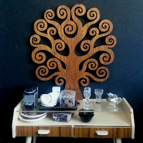 Modern miniature credenza in front of a black wall with a large wooden tree of life on it. On the credenza is a Nespresso machine, tray with cups, saucers, milk and a container of biscuits, next to two wine glasses, a water glass and a silver milk jug.