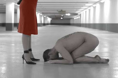 Naked man kneeling before his mistress, a woman dresses in a red dress, in an empty parking lot